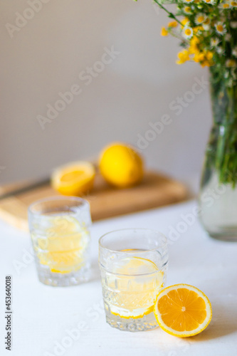 Homemade refreshing summer lemonade drink with lemon slices and ice in glasses with flowers bouquet in a vase on background