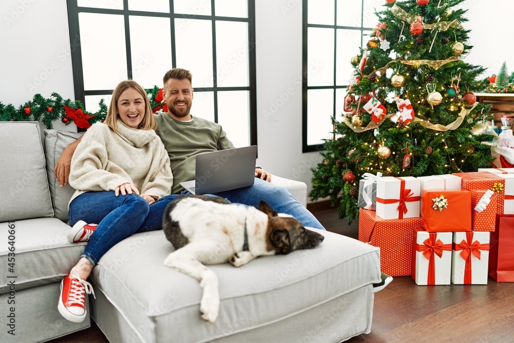 Young couple using laptop sitting by christmas tree winking looking at the camera with sexy expression, cheerful and happy face. Young couple using laptop sitting by christmas tree winking looking at the camera with sexy expression, cheerful and happy face.