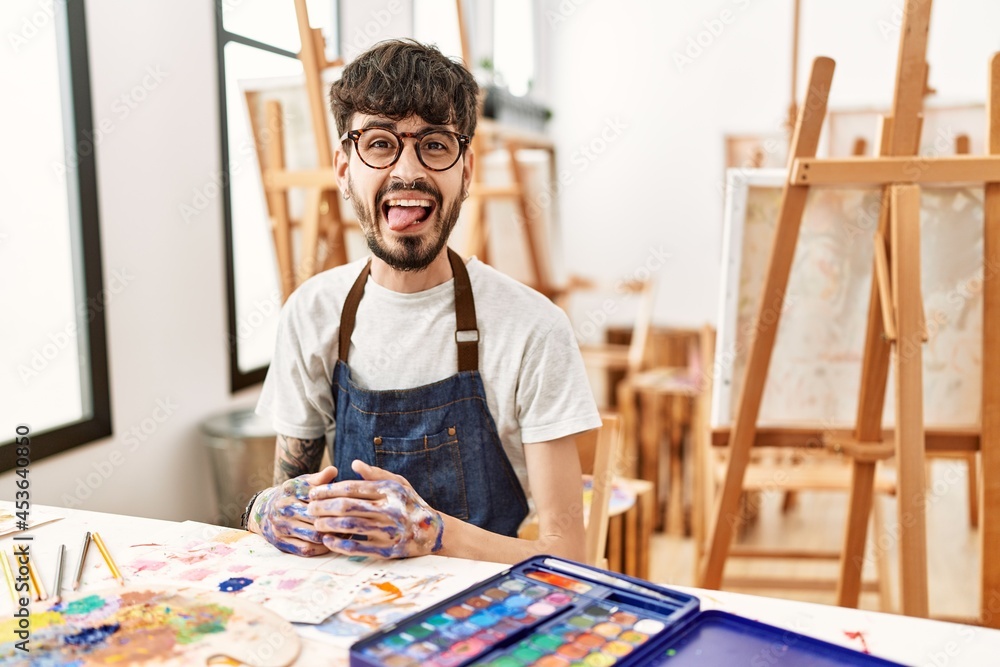 Hispanic man with beard at art studio sticking tongue out happy with funny expression. emotion concept.
