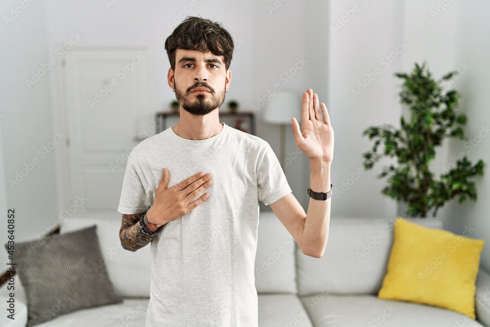 Hispanic man with beard at the living room at home swearing with hand on chest and open palm, making a loyalty promise oath Hispanic man with beard at the living room at home swearing with hand on chest and open palm, making a loyalty promise oath