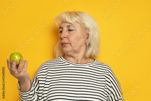 Cheerful old blonde haired woman hold a green apple on yellow background