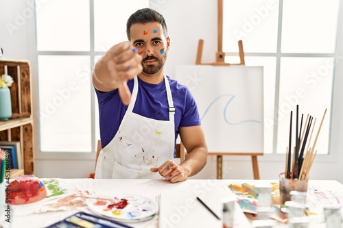 Young hispanic man with beard at art studio with painted face looking unhappy and angry showing rejection and negative with thumbs down gesture. bad expression.