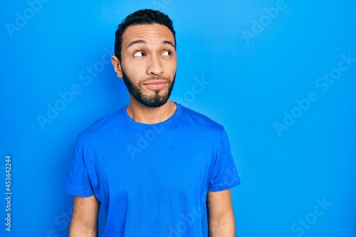 Hispanic man with beard wearing casual blue t shirt smiling looking to the side and staring away thinking.