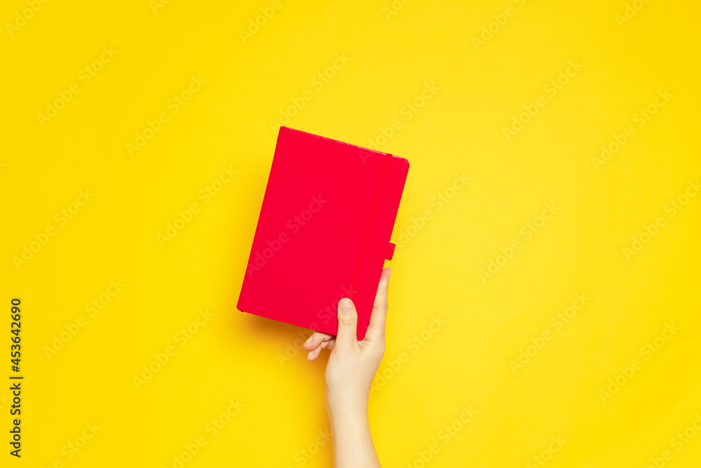 Female hand holds a red notebook for scrapes on a yellow background Female hand holds a red notebook for scrapes on a yellow background