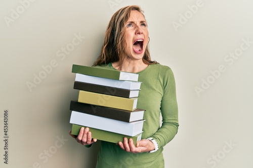 Young blonde woman holding a pile of books angry and mad screaming frustrated and furious, shouting with anger looking up.