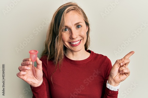 Young blonde woman holding menstrual cup smiling happy pointing with hand and finger to the side