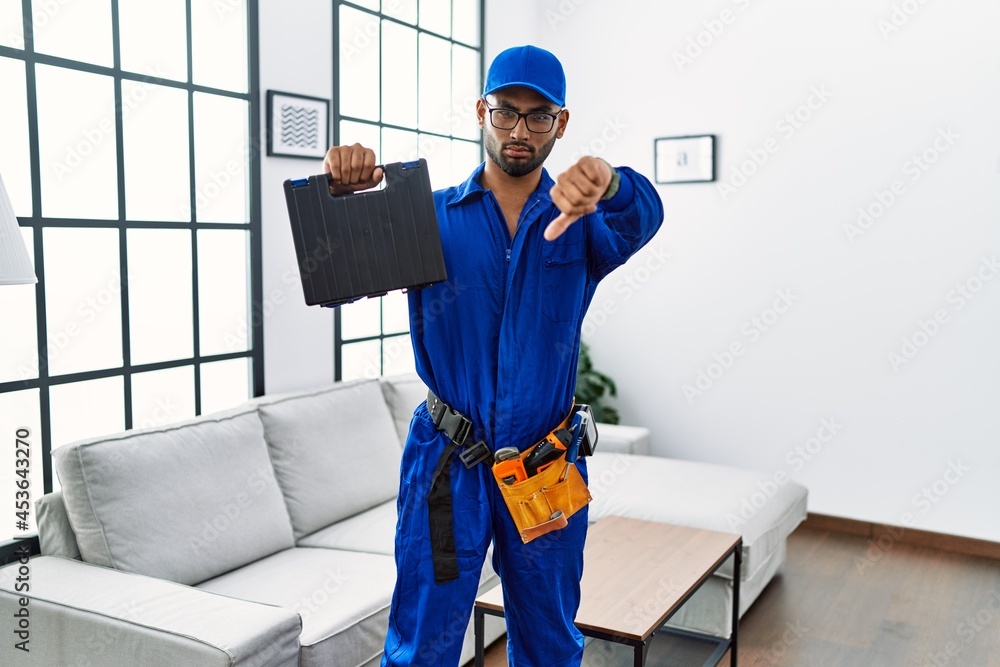 Young indian technician holding toolbox and screwdriver at house with angry face, negative sign showing dislike with thumbs down, rejection concept Young indian technician holding toolbox and screwdriver at house with angry face, negative sign showing dislike with thumbs down, rejection concept