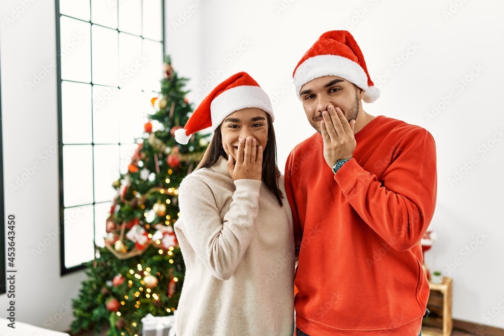 Young hispanic couple standing by christmas tree laughing and embarrassed giggle covering mouth with hands, gossip and scandal concept Young hispanic couple standing by christmas tree laughing and embarrassed giggle covering mouth with hands, gossip and scandal concept