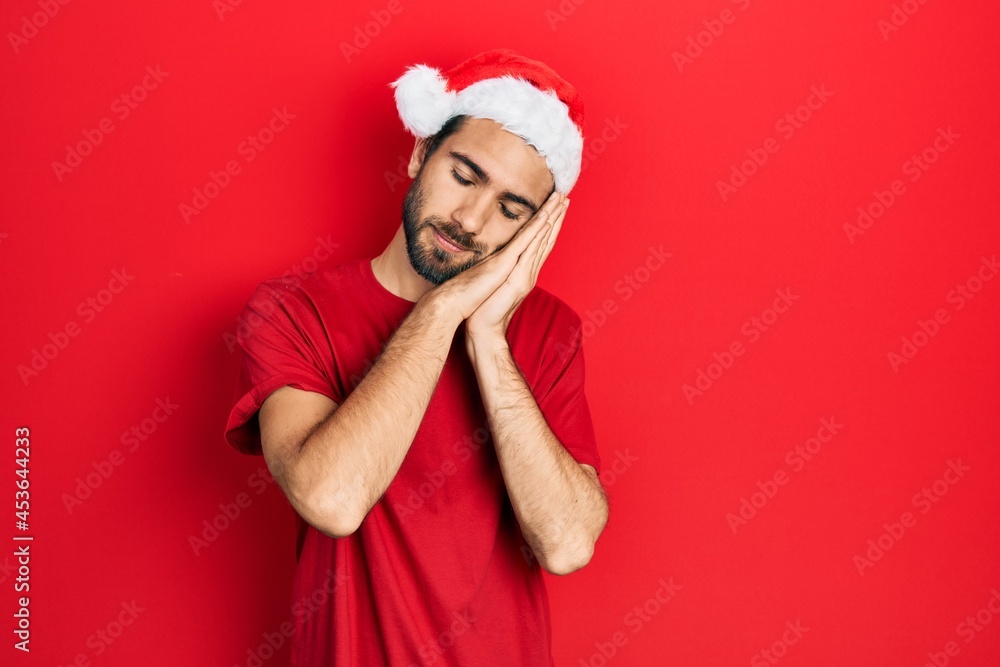 Young hispanic man wearing christmas hat sleeping tired dreaming and posing with hands together while smiling with closed eyes.