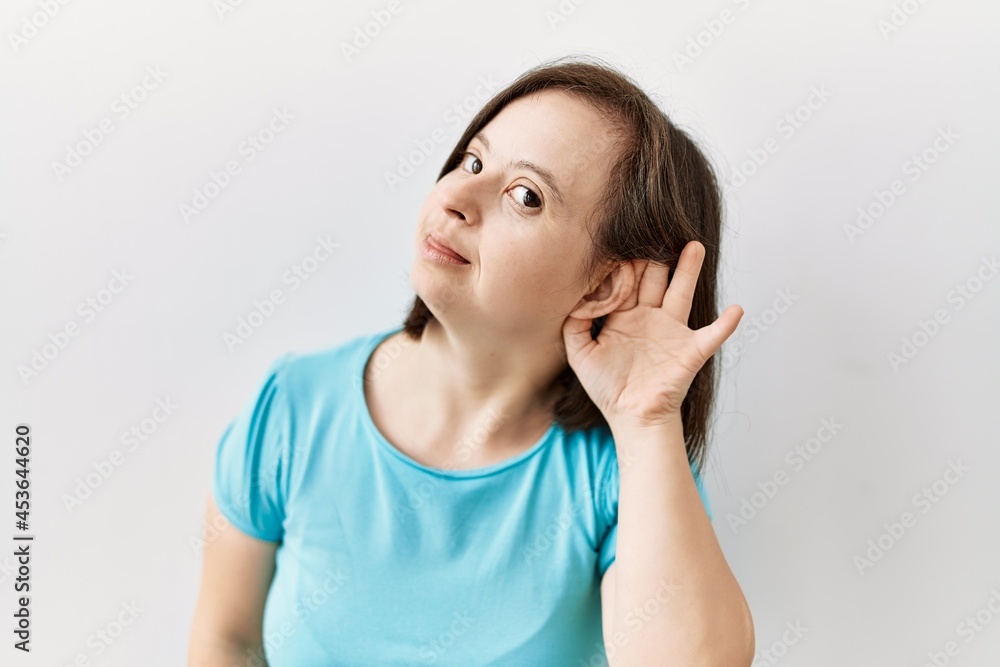 Young down syndrome woman standing over isolated background smiling with hand over ear listening an hearing to rumor or gossip. deafness concept. Young down syndrome woman standing over isolated background smiling with hand over ear listening an hearing to rumor or gossip. deafness concept.
