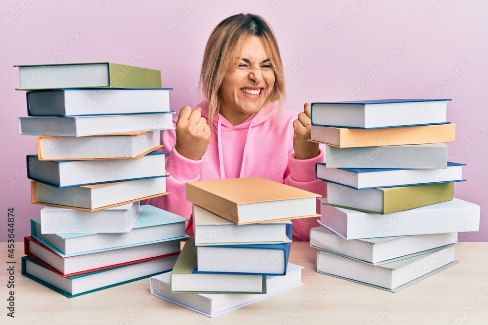 Young caucasian woman sitting on the table with books very happy and excited doing winner gesture with arms raised, smiling and screaming for success. celebration concept. Young caucasian woman sitting on the table with books very happy and excited doing winner gesture with arms raised, smiling and screaming for success. celebration concept.