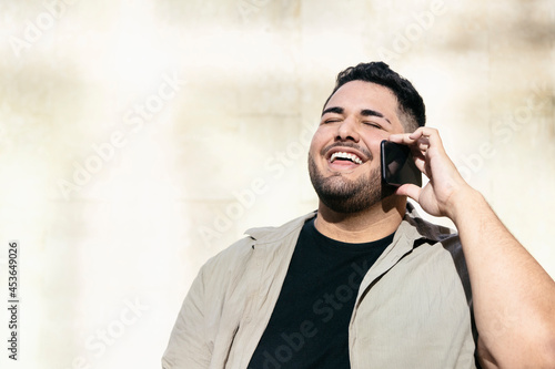 Fat latin man, talking on his cell phone, with a big smile. With a white wall of the street in the background. Latins, smartphones, business and curvy models concept. Copyspace