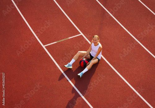 young woman is sitting in the stadium and holding a sports ball for fitness, looking at the camera. the concept of sports training, fitness, healthy lifestyle.