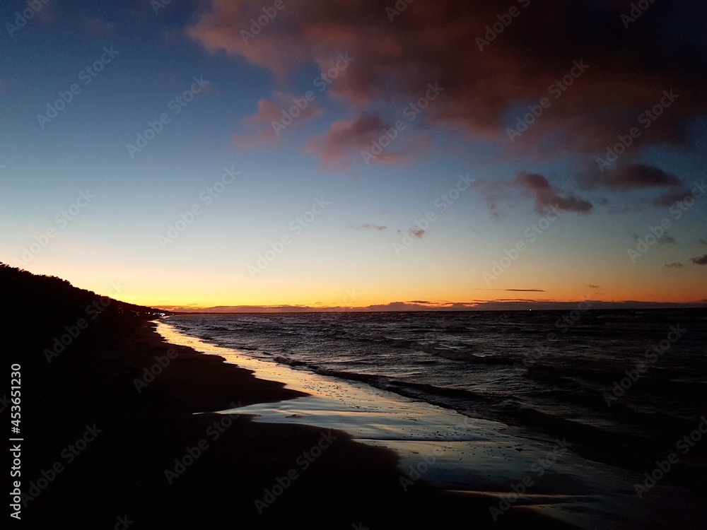 Fototapeta premium Small wave on the surface of the sea and a dark sandy beach against the blue sky after sunset