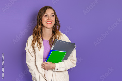 Caucasian student girl holding a book, notebook smiling, looking at the camera isolated on lilac background in studio. copy space