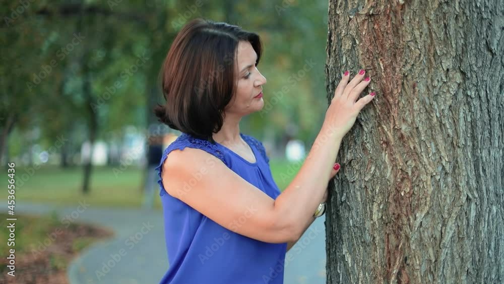 A beautiful brunette girl is standing in the park near an old big tree. She strokes the bark with her hand. The youth of a lady and the wisdom of a tree in one frame. Tenderness and severity. A beautiful brunette girl is standing in the park near an old big tree. She strokes the bark with her hand. The youth of a lady and the wisdom of a tree in one frame. Tenderness and severity.