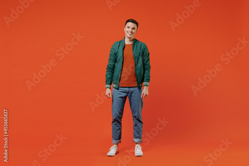 Full size body length smiling vivid happy young brunet man 20s wear red t-shirt green jacket stand look camera isolated on plain orange background studio portrait. People emotions lifestyle concept