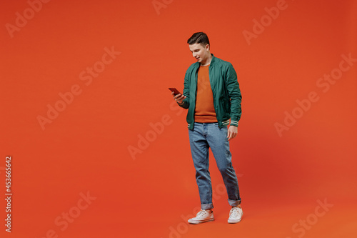 Full size body length smiling vivid happy young brunet man 20s wears red t-shirt green jacket hold in hand use mobile cell phone go step strolling isolated on plain orange background studio portrait