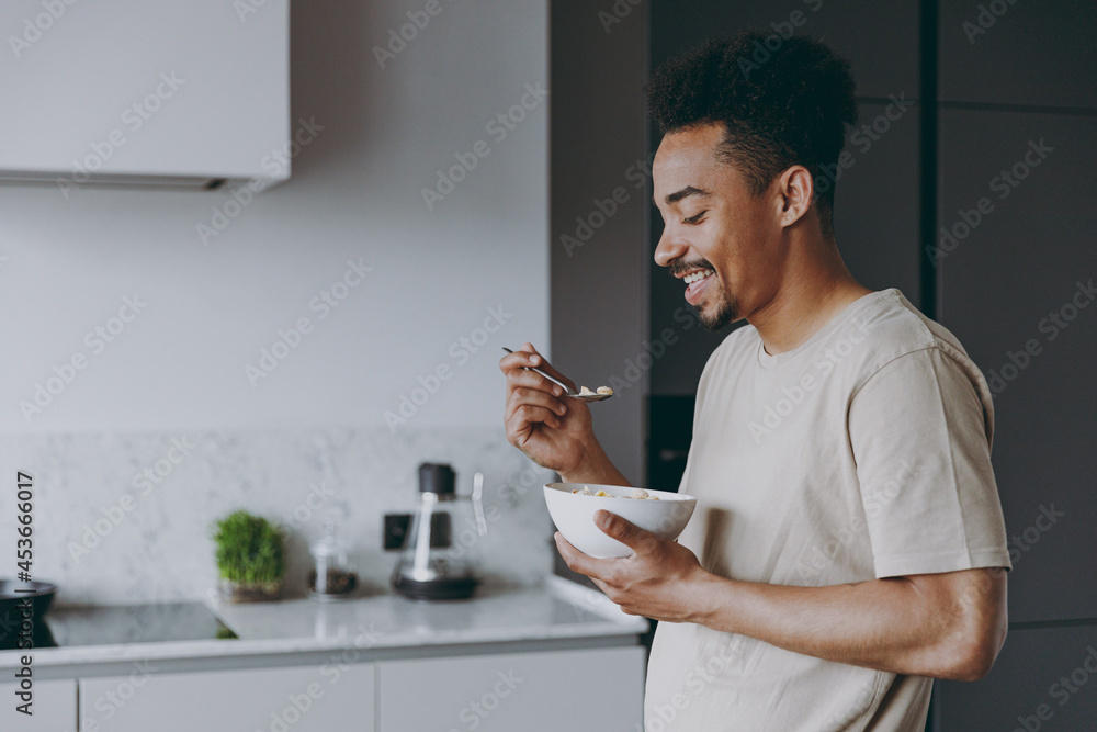 Side view young happy smiling african american man in casual clothes eat breakfast muesli cereals with milk fruit in bowl prepare cook food in light kitchen at home alone indoor. Healthy diet concept