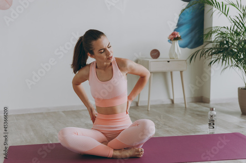 Full size young sick sporty athletic fitness trainer instructor woman in pink tracksuit do yoga exercises feel bad pain spasm small of back sprain neck position sit on mat floor at home gym inside