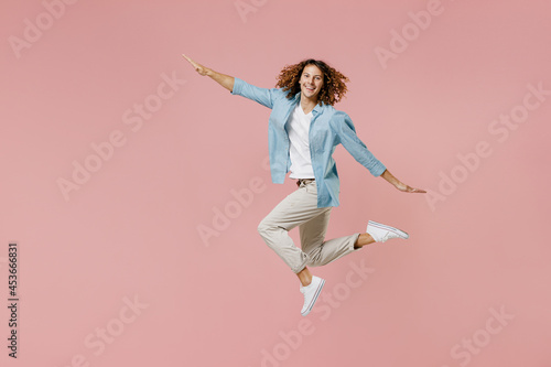Full length young excited happy carefree student fun man with long curly hair wear blue shirt white t-shirt with outstretched arms jump high isolated on pastel plain pink color wall background studio