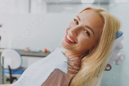 Fotografi Close up young smiling happy calm blonde woman covered by napkin sit at dental office chair indoor light modern cabinet waiting stomatologist for oral procedure