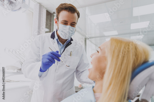 Young fun man doctor in white gown sterile mask make oral examination for patient woman sit at dentist office chair indoor cabinet near stomatologist wait for procedure Healthcare enamel treatment