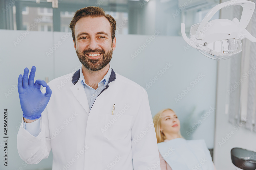 Young confident smiling happy male man doctor show ok okay gesture stand near patient woman sitting at dentist office chair indoor light cabinet near stomatologist Healthcare oral enamel treatment. Young confident smiling happy male man doctor show ok okay gesture stand near patient woman sitting at dentist office chair indoor light cabinet near stomatologist Healthcare oral enamel treatment.