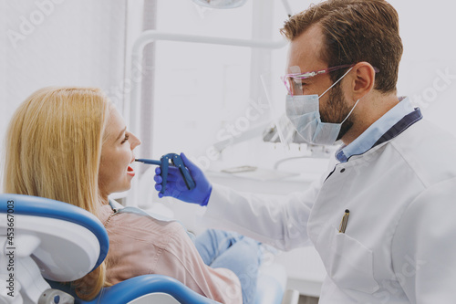 Young caucasian man doctor in face shields and googles hold dental equipment instrument device examine mouth cavity of patient woman sitting at dentist office chair indoor cabinet near stomatologist.