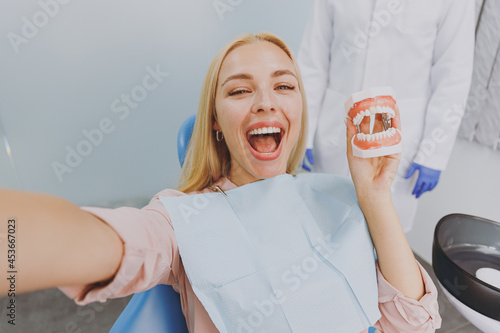 Close up young excited woman do selfie shot pov on mobile phone hold artificial jaw sit at dental office chair indoor cabinet wait stomatologist for oral procedure Healthcare orthodontal treatment. photo