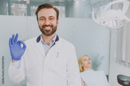 Young confident smiling happy male man doctor show ok okay gesture stand near patient woman sitting at dentist office chair indoor light cabinet near stomatologist Healthcare oral enamel treatment.