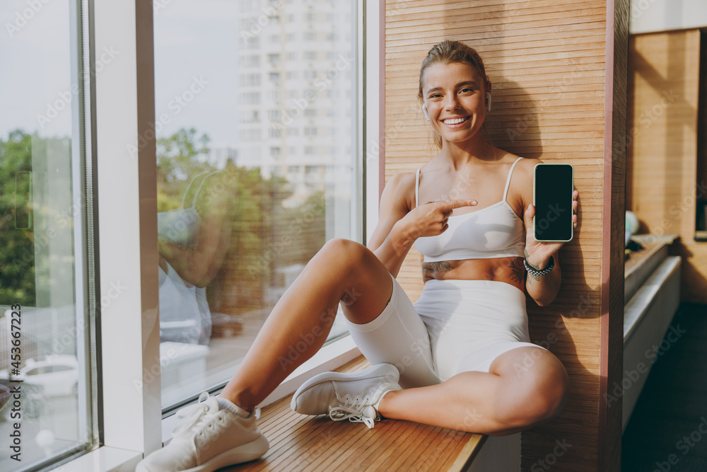 Fun young skinny sportswoman woman in white sportswear earphones listen music point finger on mobile cell phone with blank screen area sit on windowsill of gym rest indoor People lifestyle concept