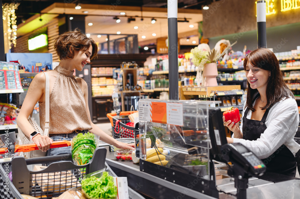 Young cheerful happy woman 20s wear casual clothes shopping at supermaket with grocery cart stand at store checkout pays for groceries cashier inside hypermarket Purchasing gastronomy food concept