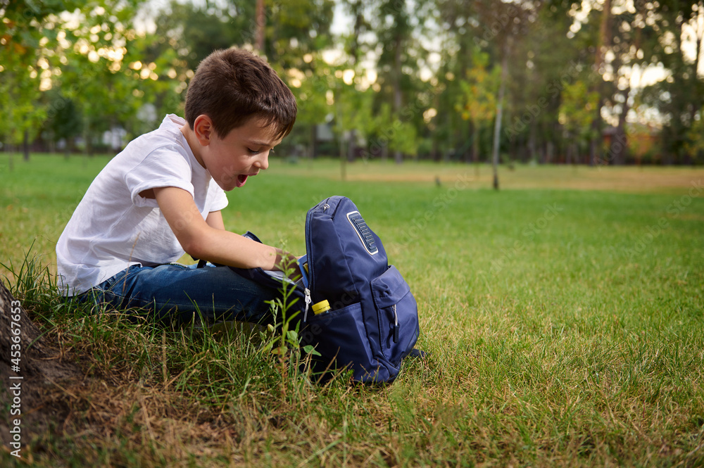 Elementary aged school boy in casual denim resting on green grass after his first day at school. Adorable child with school bag during recreation on beautiful sunny day. Back to school concept Elementary aged school boy in casual denim resting on green grass after his first day at school. Adorable child with school bag during recreation on beautiful sunny day. Back to school concept