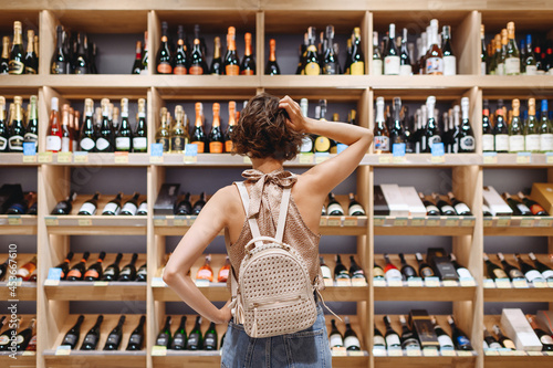 Murais de parede Back view puzzled minded young woman in casual clothes shopping at supermaket grocery store buy choosing wine alcohol bottle scratch head inside hypermarket