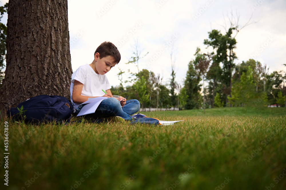 Serene child schoolboy concentrating on doing homework, sitting on the city park green grass, enjoying fresh air during his recreation after first day at school. Back to school concept. September. Serene child schoolboy concentrating on doing homework, sitting on the city park green grass, enjoying fresh air during his recreation after first day at school. Back to school concept. September.