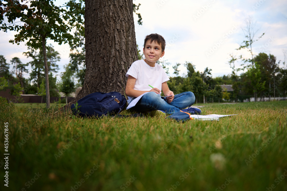 Serene handsome school boy doing homework, sitting on city park green grass, looking to the side, enjoying fresh air during his recreation after first day at school. Back to school concept. September. Serene handsome school boy doing homework, sitting on city park green grass, looking to the side, enjoying fresh air during his recreation after first day at school. Back to school concept. September.