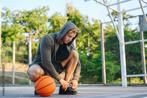 Young sporty caucasian sportsman man 20s wearing grey sportswear hood looking aside training sit laces up sneakers shoes with ball at basketball game playground court Outdoor courtyard sports concept