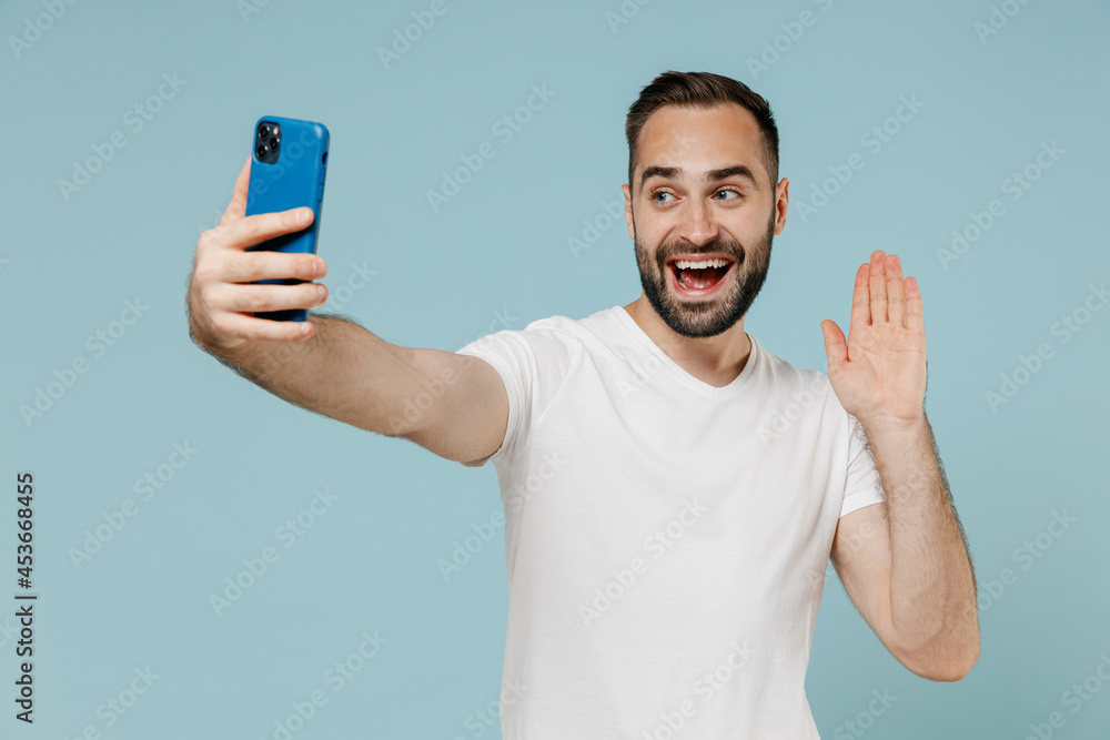 Young smiling happy man in white t-shirt doing selfie shot on mobile cell phone post photo on social network waving hand talk by video call isolated on plain pastel light blue color background studio Young smiling happy man in white t-shirt doing selfie shot on mobile cell phone post photo on social network waving hand talk by video call isolated on plain pastel light blue color background studio
