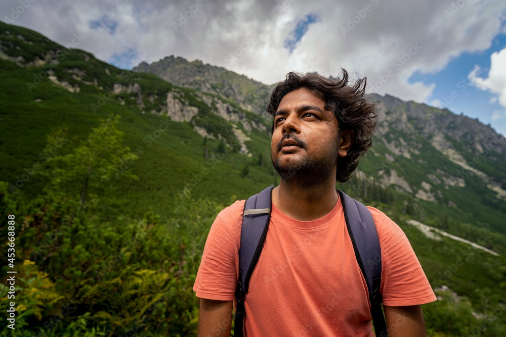 Portrait of a focused adventurer Indian tourist man looking up towards the way forward with bag pack against mountains during hike. Summer adventure and sports concept