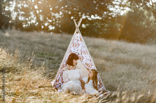 Summer holidays. Pretty young mother and her cute little daughter spending time together outdoors. Mom and little girl wearing white dress, boho feather heair accessories and sitting near teepee tent photo