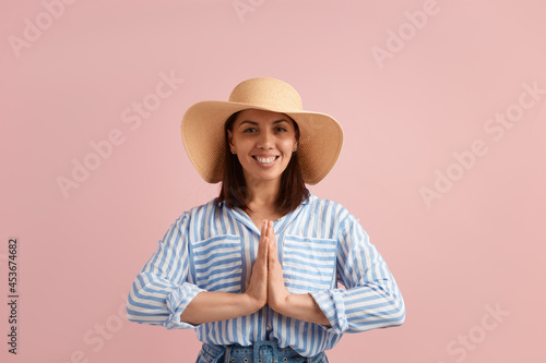 Smiling woman with dark hair practices yoga, makes namaste gesture, prays, is grateful, feels harmony, wears straw hat, striped shirt, jeans with belt, on pink background. Summer emotions concept.
