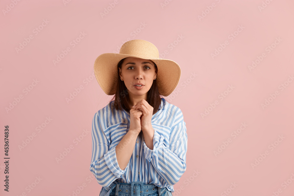 Worried woman with dark hair folds hands over chest, hopes she will receive bonus, go on vacation, wears straw hat, striped shirt, jeans with belt, on pink background. Summer emotions concept. Worried woman with dark hair folds hands over chest, hopes she will receive bonus, go on vacation, wears straw hat, striped shirt, jeans with belt, on pink background. Summer emotions concept.