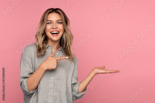 Excited young woman holding palm open for new product or sale presentation. Overjoyed happy blond girl with open mouth point finger at arm with copy space for promotion isolated over pink studio wall photo