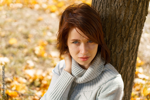 A young woman in a gray sweater sits alone, leaning against a tree on a beautiful day in the park. Golden autumn. Thinking about life. Spending time alone in nature. Calming atmosphere.