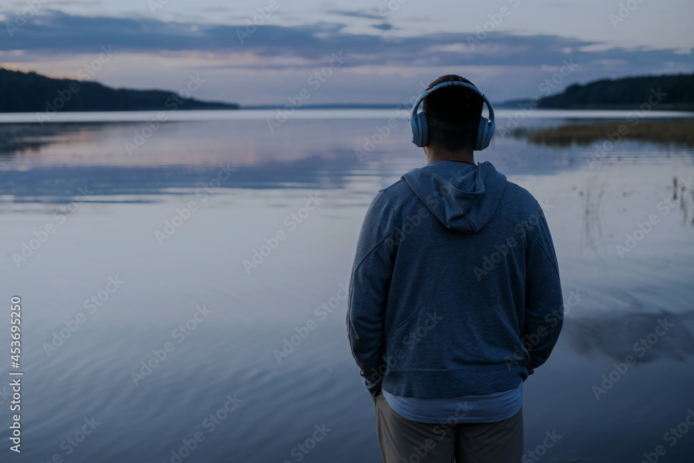 A young man is listening to an audiobook with wireless headphones. Beautiful view of the lake and the sunset.