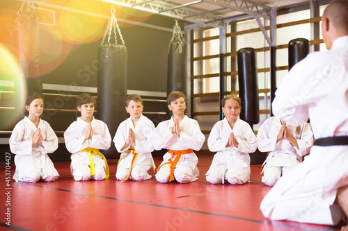 Group of karate kids in white kimono kneeling in front of their trainer on gym floor.