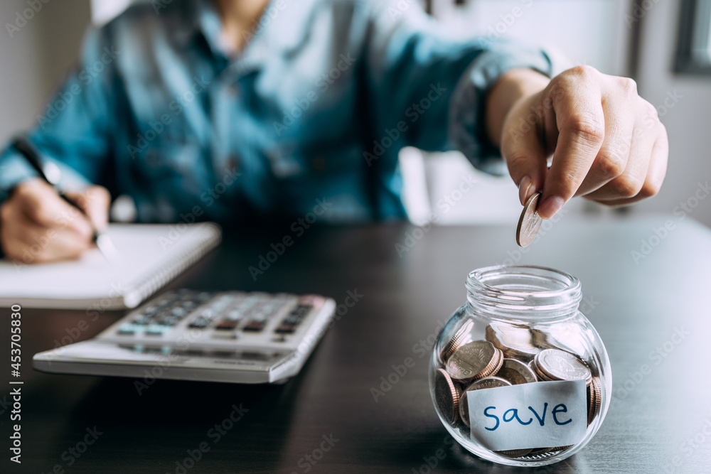 Fototapeta premium Close up of hand young women insert coins in jug glass with doing Household income and expenses account, insurance, retirement fund, Investment and Saving Money concept.