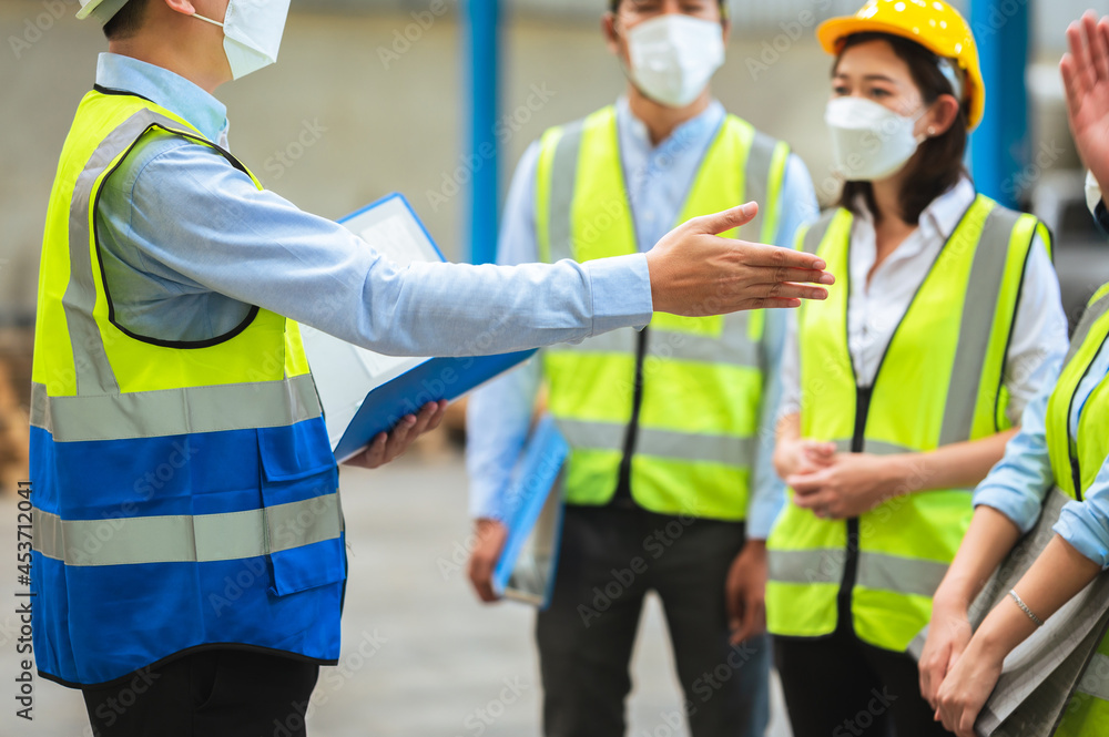 The factory employee consists of engineers, foreman, technicians, and related department staff. Wear a mask, hard hat, and vest. meeting before starting work inside the warehouse. Teamwork concept. The factory employee consists of engineers, foreman, technicians, and related department staff. Wear a mask, hard hat, and vest. meeting before starting work inside the warehouse. Teamwork concept.