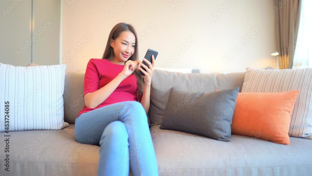 Asian woman using a mobile phone while lying on sofa in her living room. Happy female using smartphone for texting, reading, watching video and surfing online at home. Lifestyle woman home concept Asian woman using a mobile phone while lying on sofa in her living room. Happy female using smartphone for texting, reading, watching video and surfing online at home. Lifestyle woman home concept